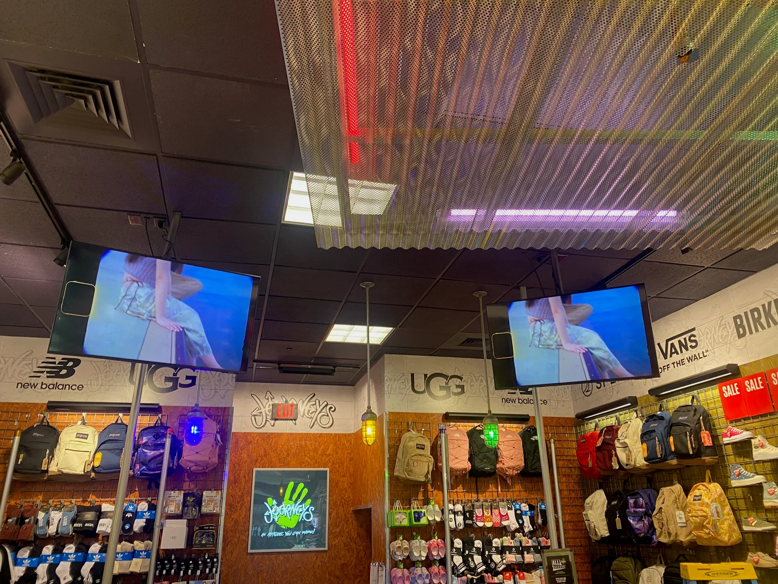 Two ceiling-mounted screens showing the same video above footwear and backpack displays at Journeys.