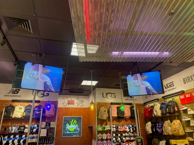 Two ceiling-mounted screens showing the same video above footwear and backpack displays at Journeys.