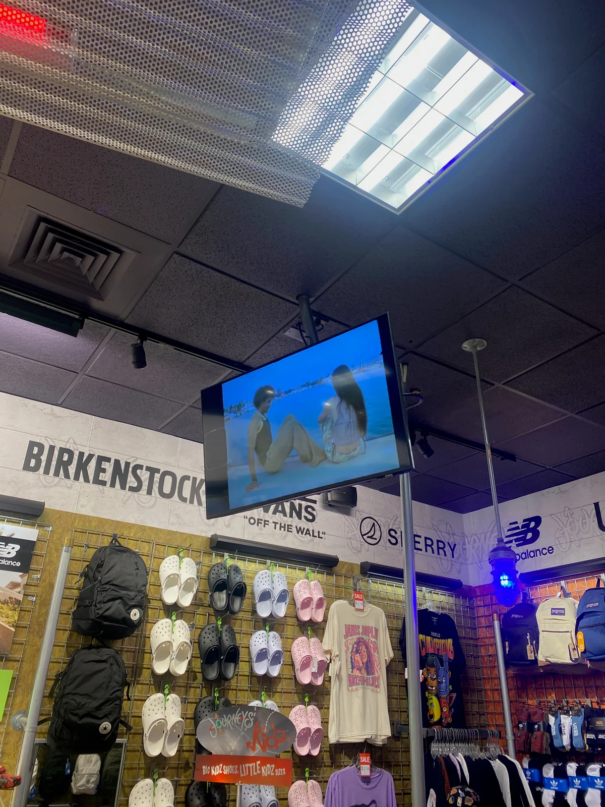 A retail video screen suspended below the drop ceiling, viewed from below beside footwear displays.